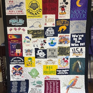 Peut inclure: Une courtepointe souvenir rectangulaire composée de divers panneaux de t-shirts colorés présentant des impressions graphiques variées, incluant des personnages de dessins animés, des logos sportifs, du texte et des illustrations, le tout assemblé avec un motif matelassé blanc visible.