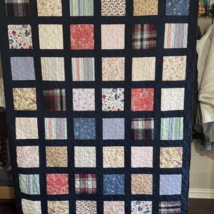 Könnte beinhalten: Eine marineblaue Steppdecke mit Patchwork-Muster. Die Steppdecke besteht aus Quadraten aus verschiedenen Stoffen, die jeweils ein einzigartiges Muster aufweisen. Die Steppdecke hängt an einem Gestell.