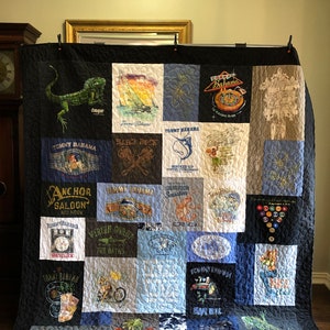 Peut inclure: Une courtepointe faite de divers t-shirts, présentant un mélange de couleurs bleues, noires, blanches et rouges. La courtepointe est composée de carrés, chacun avec un motif différent. Certains des motifs comprennent un palmier, un bateau, un poisson et un phare. La courtepointe est signée "Larry Torrence".
