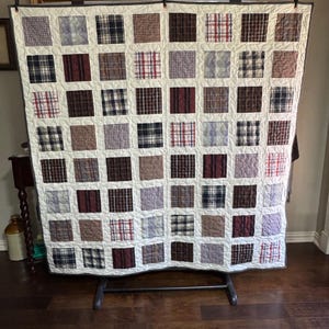 May include: A patchwork quilt with a variety of plaid squares in shades of brown, red, blue, and purple, set against a white background. The quilt is displayed on a metal stand. The quilt is handmade.