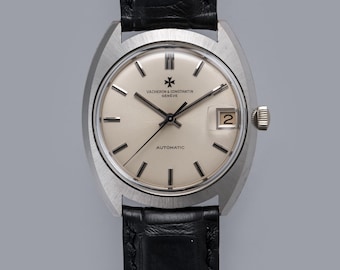 May include: A Vacheron & Constantin Genève automatic wristwatch with a silver dial, silver-tone baton indices, and a date window at the three o'clock position. The watch features a brushed stainless steel case and a black leather strap.