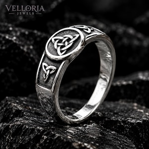 May include: A silver-toned ring featuring a circular centerpiece with an embossed triquetra knot design. The band includes additional smaller triquetra symbols on the sides, with a dark, oxidized finish in the recessed areas of the metal.