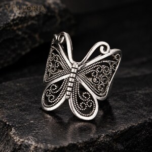 May include: A silver-toned ring shaped like a butterfly with intricate filigree cutouts and a textured, oxidized finish. The wings feature detailed patterns of small dots and swirling lines, with the text Velloria Jewels visible in the upper corner.