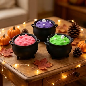 May include: Three black miniature cauldrons with rounded bodies and small handles, each filled with a smooth, bubbly substance in bright pink, vibrant purple, and lime green, featuring small white accents on the surface of the liquid.