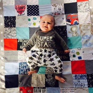 Könnte beinhalten: Ein Baby in einem grauen Strampler mit dem Aufdruck "SUPER STAR" und einer Hose mit Sternenmuster liegt auf einer Patchwork-Decke. Die Decke besteht aus verschiedenen Stoffquadraten mit Text und Bildern, darunter "I love mummy daddy!" und eine Mickey Mouse Grafik.
