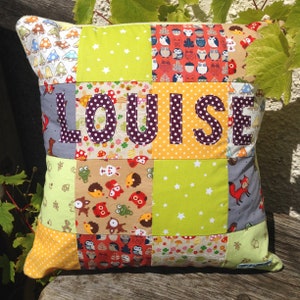 May include: A patchwork pillow with the name "LOUISE" in large, purple polka-dot letters. The pillow features various fabric squares with patterns like mushrooms, owls, stars, and foxes. The color palette includes green, orange, yellow, and gray.