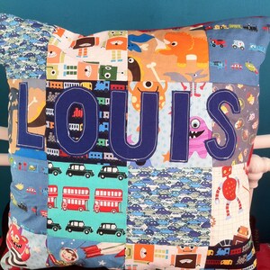 Op de afbeelding: Een patchwork kussen met de naam "LOUIS" in marineblauw aangebracht. Het kussen heeft verschillende stoffen vierkanten met patronen van auto's, monsters, robots en treinen. De achtergrond is een blauwgroene kleur.