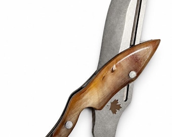 Peut inclure: Un couteau pliant avec une lame métallique dotée d'une fente et d'un petit emblème de feuille d'érable marron. Le manche est composé de deux pièces de corne polie brun clair au grain naturel visible, fixées par des rivets en métal argenté.