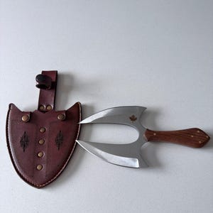 Peut inclure: Un couteau de lancer à double lame avec un manche en bois et un petit emblème de feuille d'érable sur la lame, accompagné d'un étui en cuir marron foncé avec des coutures décoratives, des rivets métalliques et une boucle de ceinture.
