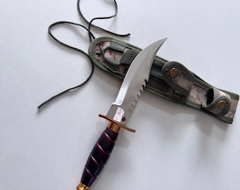 Peut inclure: Un couteau à lame fixe avec un dos dentelé, une lame argentée et un manche rainuré marron foncé doté d'une garde dorée. Le couteau porte une petite étiquette blanche avec du texte et est accompagné d'un étui en tissu à motif camouflage.