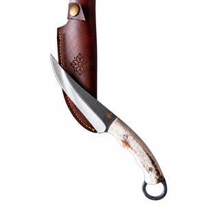 May include: A fixed-blade knife with a curved silver steel blade, a textured bone handle, and a metal ring at the pommel. A small maple leaf icon is etched on the blade. It is paired with a dark brown leather sheath featuring a brass snap.