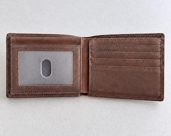 Peut inclure: Un portefeuille bifold en cuir marron présenté en position ouverte. Le côté gauche comporte une fenêtre transparente pour carte d'identité avec une découpe pour le pouce, tandis que le côté droit contient plusieurs fentes pour cartes.