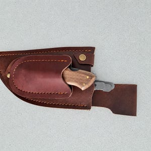 Peut inclure: Un étui en cuir marron avec des coutures orange et un bouton-pression en laiton, contenant un couteau pliant avec un manche en bois marron clair et une lame en métal argenté, ainsi que deux lames en métal argenté supplémentaires.