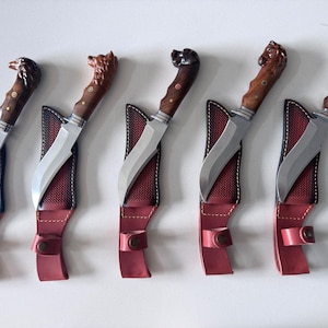 May include: Five fixed-blade knives with silver-toned steel blades and carved wooden handles shaped like animal heads. Each knife is paired with a red and black textured leather sheath featuring a snap closure and visible stitching details.