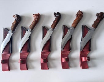 Peut inclure: Cinq couteaux à lame fixe avec des lames en acier argenté et des manches en bois sculpté en forme de têtes d'animaux. Chaque couteau est accompagné d'un étui en cuir texturé rouge et noir avec une fermeture à pression et des coutures.