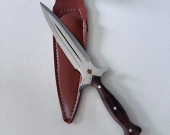 Peut inclure: Un poignard à double tranchant avec une lame en acier argenté arborant un petit emblème de feuille d'érable près de la garde. Le manche est en bois brun foncé avec trois rivets argentés. Il est accompagné d'un étui en cuir brun.