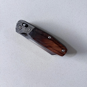 May include: A folding pocket knife featuring a dark brown wood handle with a visible grain pattern. The bolster is made of silver-toned metal with an intricate floral scrollwork engraving. The knife is secured with three small silver-toned screws.