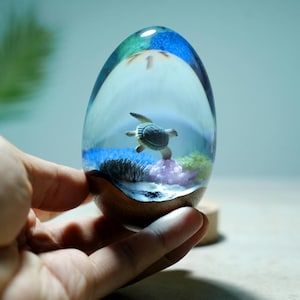 May include: An egg-shaped resin sculpture featuring a miniature sea turtle swimming above a colorful underwater scene with pink coral, blue sand, and green seaweed, set within a clear, polished resin dome atop a natural wooden base.