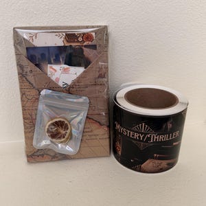 May include: A rectangular package wrapped in vintage map paper containing a small clear pouch with a dried citrus slice, alongside a roll of black adhesive labels featuring the text Mystery/Thriller and an illustration of a book and glass.