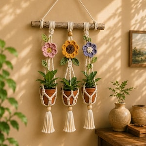 May include: A macrame wall hanging featuring three terracotta pots suspended by off-white cotton cord from a wooden branch. Each pot holds a green plant and is decorated with a crocheted flower in pink, yellow, or purple, plus green leaves.