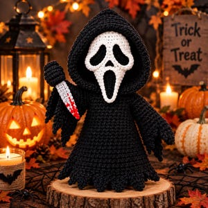 May include: A crocheted figure wearing a black hooded robe and a white mask with elongated eye and mouth openings. The figure holds a small white knife with red blood-like stains on the blade. The item is crafted with visible yarn stitches.