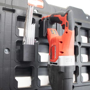 May include: A red and black reciprocating saw mounted on a black plastic wall organizer panel. A black plastic holder attached to the panel contains several metal saw blades. The tool features a textured grip, a trigger, and a battery housing.
