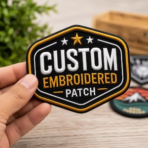 Puede incluir: Un parche bordado hexagonal con fondo negro y borde dorado. El texto CUSTOM está en blanco, EMBROIDERED en dorado y PATCH en blanco. Tres estrellas están centradas sobre el texto, una estrella dorada flanqueada por dos estrellas blancas.