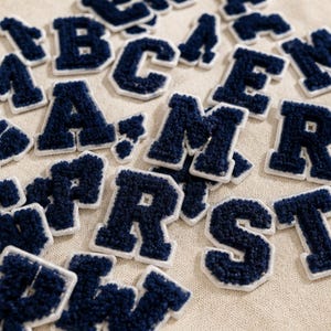May include: A collection of navy blue varsity-style letter patches featuring a textured, fuzzy chenille surface. Each individual letter is outlined with a white border, creating a bold, collegiate appearance suitable for various textile applications.