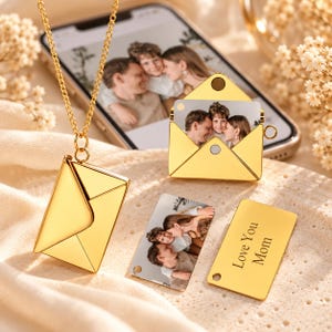 Puede incluir: Un collar con colgante en forma de sobre de metal dorado y cadena a juego. El relicario se abre para mostrar una placa metálica rectangular con una foto familiar impresa. Una segunda placa metálica lleva grabado el texto Love You Mom.