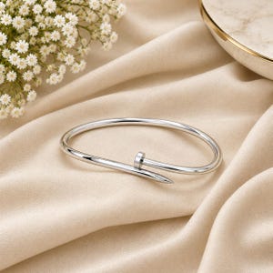 May include: A polished silver-tone bangle bracelet shaped like a bent nail. The design features a rounded head at one end and a tapered point at the other, with the two ends overlapping to form a continuous, circular loop for the wrist.