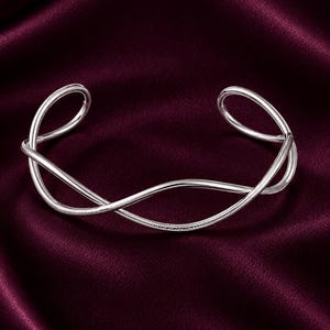May include: A silver-toned open cuff bracelet featuring a fluid, intertwined wave design. The metal surface displays a smooth, polished finish on the upper curves and a fine, textured cross-hatch pattern along the lower crossing sections.