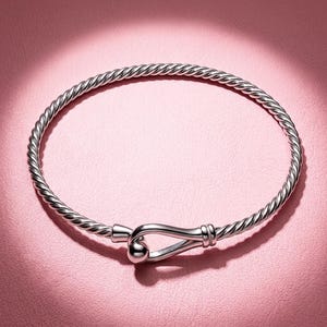 Può includere: Un bracciale rigido color argento caratterizzato da una fascia a corda intrecciata e un meccanismo di chiusura a gancio. La chiusura è composta da un gancio liscio e arrotondato che si inserisce in una piccola sfera lucida in metallo.