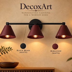 Könnte beinhalten: Eine dreiflammige Wandleuchte mit einer mattschwarzen horizontalen Stange und runder Wandhalterung. Sie besitzt drei konische, bordeauxrote matte Lampenschirme, die jeweils eine Glühbirne enthalten. Text: DecoxArt, Black Brass (Matte), Burgundy (Matte).