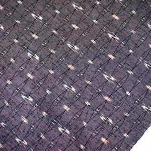 May include: Close-up of a dark grey fabric featuring a repeating pattern of white and light brown shapes. Black lines intersect the pattern diagonally. The fabric has a textured appearance and a slightly faded look.