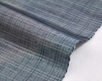 May include: Close-up of a folded piece of fabric with a plaid pattern. The fabric is a muted blue-gray color with thin white and dark blue lines creating the grid. The edge of the fabric is unfinished.
