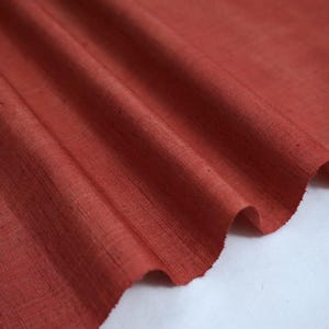 May include: Close-up of a rust-coloured fabric with a subtle texture. The fabric is folded and draped, showcasing its soft drape and natural fibres.