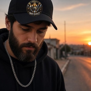 May include: A black baseball cap featuring a circular patch on the front with a gold-coloured emblem, a central letter B, and the text DEFI KINGZ. The cap has a structured crown and a curved brim, constructed from a solid black fabric material.