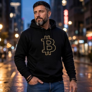 May include: A black hooded sweatshirt featuring a gold-colored Bitcoin symbol printed on the center of the chest. The garment includes a drawstring hood, long sleeves with ribbed cuffs, and a front kangaroo pocket, worn with blue denim jeans.