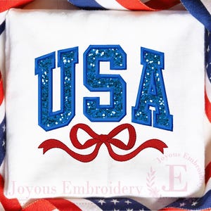 Op de afbeelding: Witte stof met het woord "USA" in blauwe glitterletters en een rode strik. De letters zijn blauw omlijnd. De afbeelding is omlijst met rood, wit en blauw lint.