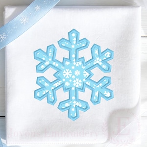 May include: A white square fabric napkin featuring a central embroidered snowflake design. The snowflake has a light blue border and a light blue interior filled with a pattern of smaller white snowflakes. A light blue ribbon with white snowflake prints is placed diagonally across the top left corner.