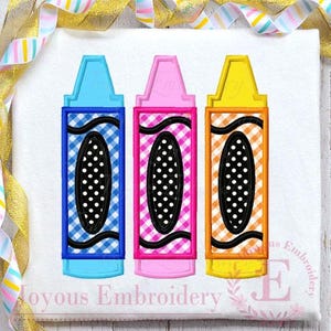 May include: Three embroidered crayon designs on a white fabric background. The left crayon is blue with a blue gingham pattern, the center is pink with a pink gingham pattern, and the right is yellow with an orange gingham pattern.