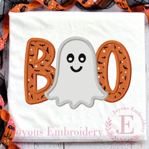 May include: White t-shirt with a Halloween-themed design. The word "BOO" is embroidered in orange with a pattern of bats and stars. A smiling ghost is centered between the letters. The shirt is decorated with orange and black ribbon.