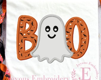 May include: White t-shirt featuring a Halloween design. The word "BOO" is embroidered in orange, patterned with bats and stars. A smiling ghost is positioned between the letters. The shirt is decorated with orange and black ribbon.