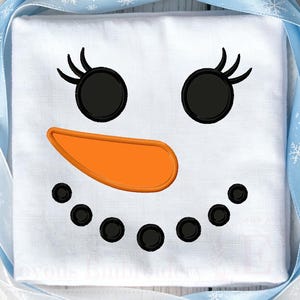 May include: A white fabric square featuring an embroidered snowman face with two black circular eyes with eyelashes, an orange carrot-shaped nose, and a curved mouth made of six small black circles, framed by a light blue ribbon with snowflakes.