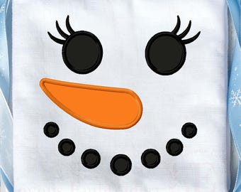 May include: A white fabric square featuring an embroidered snowman face with two black circular eyes with eyelashes, an orange carrot-shaped nose, and a curved mouth made of six small black circles, framed by a light blue ribbon with snowflakes.