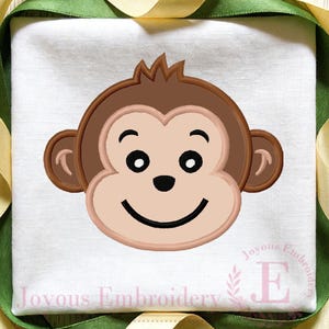 Pode incluir: Rosto de macaco bordado com uma expressão sorridente. O macaco tem pelo, orelhas e cabelo castanhos, com um rosto castanho claro e olhos e boca pretos. O texto "Joyous Embroidery" está na parte inferior.