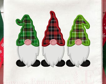 May include: A white fabric square featuring an embroidered design of three gnomes. Each gnome has a white beard, a pink nose, and black shoes. The left and right gnomes wear green plaid hats, while the center gnome wears a red plaid hat.