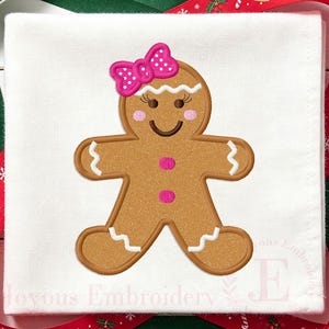 Puede incluir: Un cuadrado de tela blanca con un diseño bordado de una figura de pan de jengibre marrón. La figura tiene un lazo rosa con lunares, mejillas rosas, una boca sonriente y dos botones rosas. Detalles de glaseado blanco acentúan las muñecas, tobillos y cabeza.