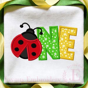 May include: White fabric with a colorful embroidered design featuring a ladybug and the word "ONE". The ladybug is red with black spots, and the letters are green and yellow.