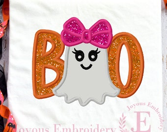May include: A white t-shirt featuring an embroidered design of the word BOO in orange glitter fabric. A light grey ghost with a bright pink glitter bow sits in the centre, replacing the letter O. The ghost has black eyes and a smiling mouth.